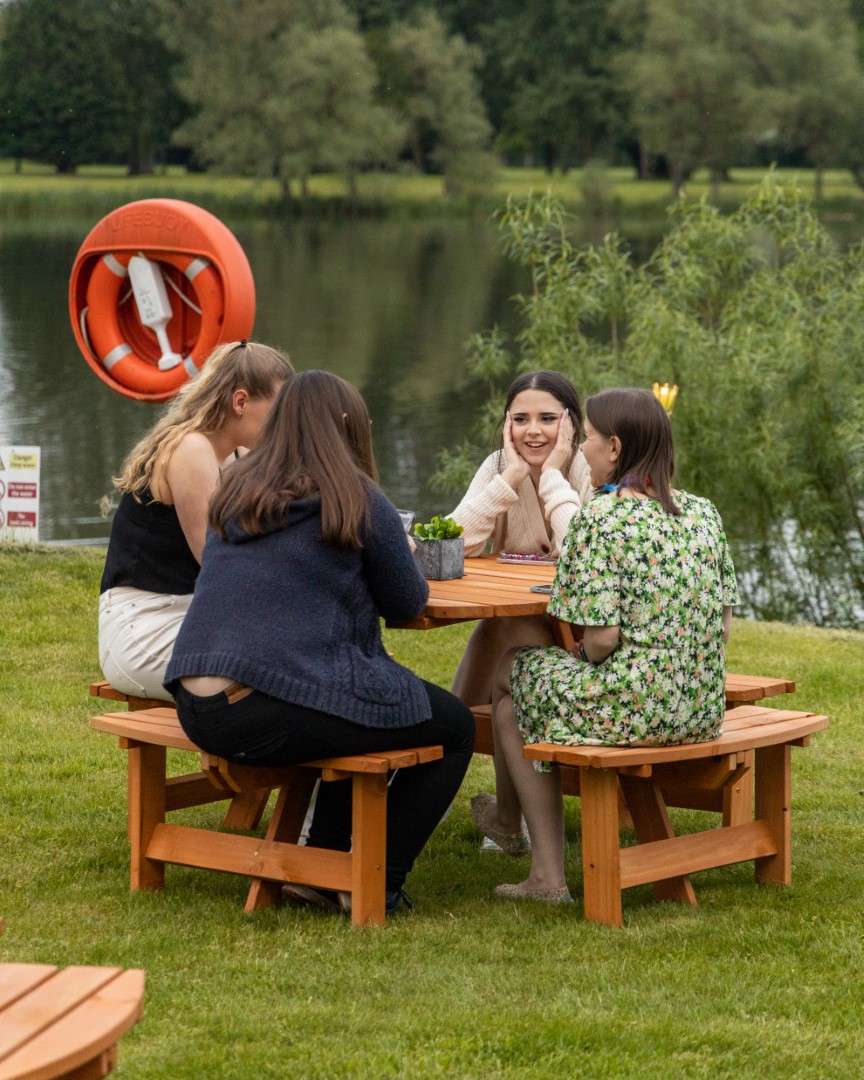 four young women sitting at wooden picnic table by lake