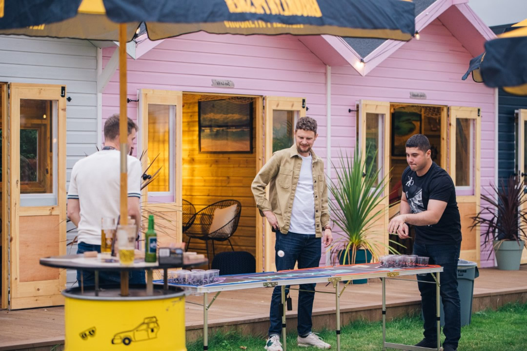 three men playing beer pong outside pink summer huts