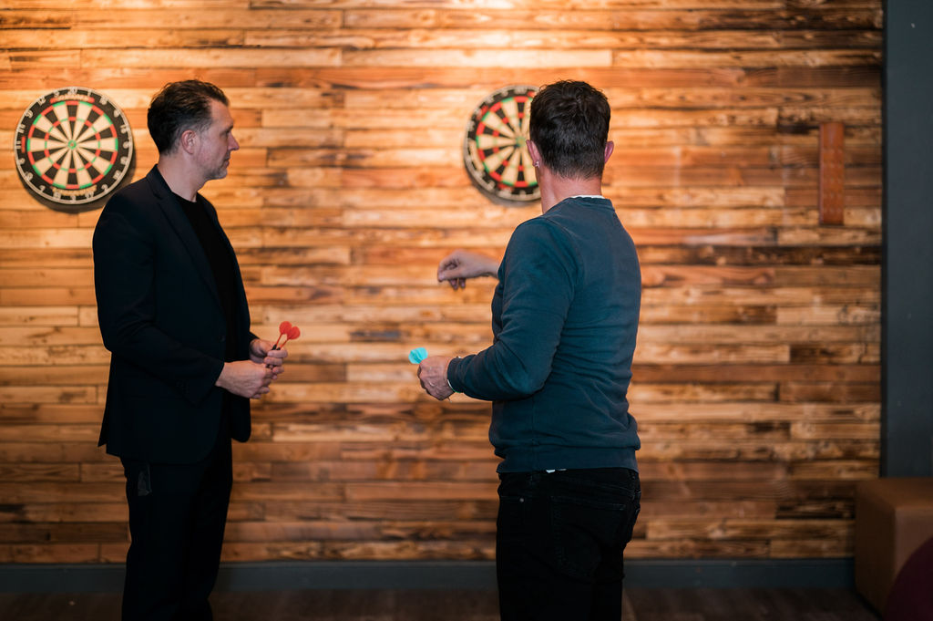two men playing darts indoors wooden wall background