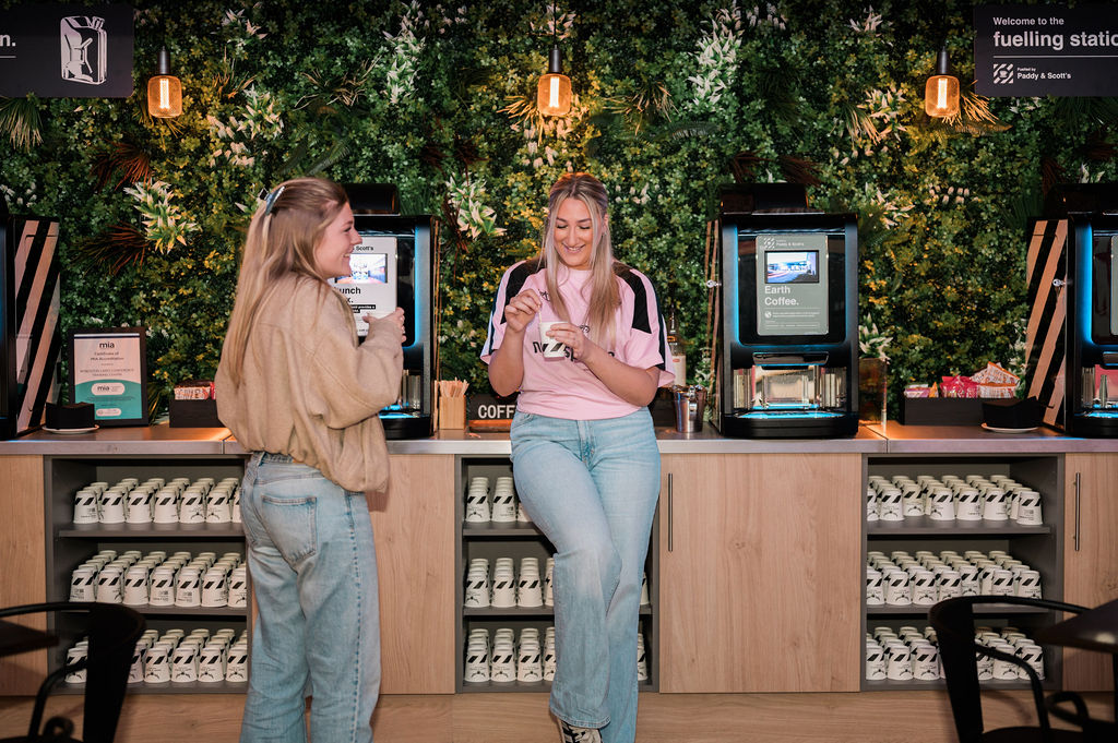 two young women chatting by coffee machines indoors